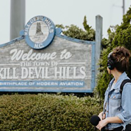 Delia D'Ambra looking at a sign that says "Welcom to the Town of Kill Devil Hills"